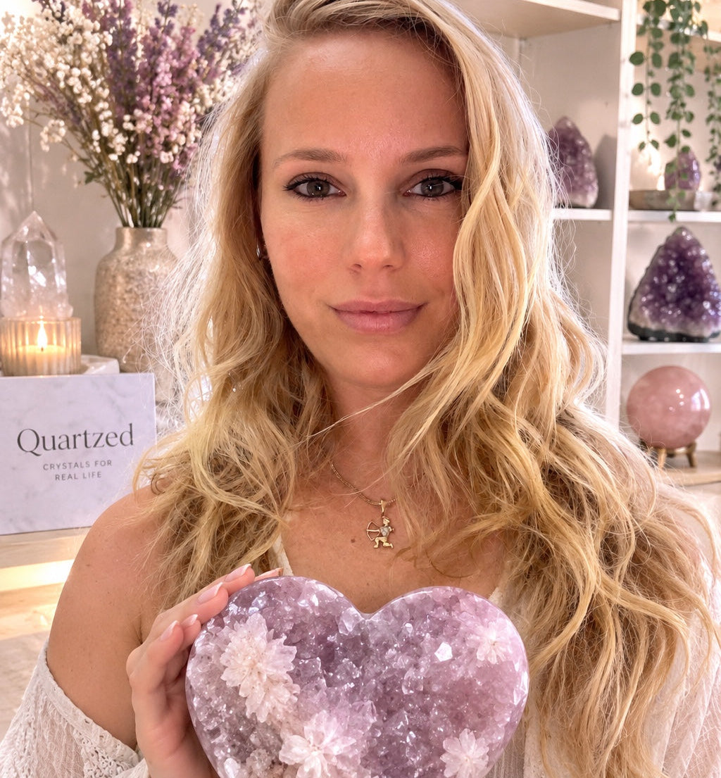 A blonde woman holds a heart-shaped amethyst crystal cluster in her hands, with a shop shelf and dried flowers in the background. The brand name 'Quartzed' is visible on a tag to the left.